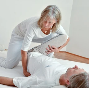 Shiatsu-Behandlung in Rückenlage, es wird mit angewinkelten Fingern von unten auf dem Kreuzbein behandelt.